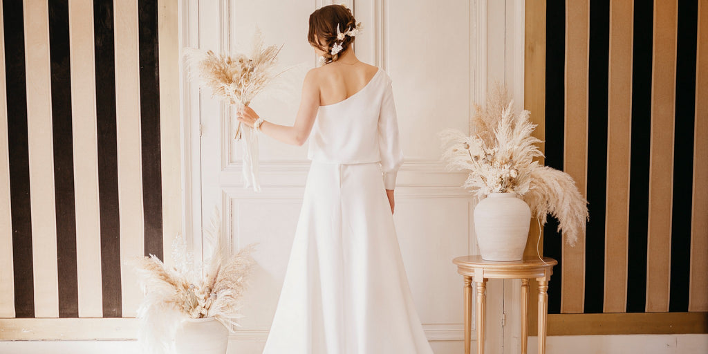 Mariée de dos en robe blanche longue accompagné de diverses créations fleuries en fleurs séchées blanches comme de la pampa